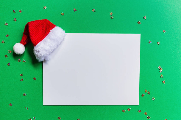 Blank of paper with red Santa hat on green background with confetti. new year concept. Greeting card, xmas celebration 2020. Flat lay, template, top view, copy space, mockup - Φωτογραφία, εικόνα