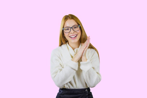 Portrait of a cute red-haired girl with glasses and a white sweater folded hands together holding them in the side smiling while opening her mouth looking at the camera on an isolated background - 写真・画像