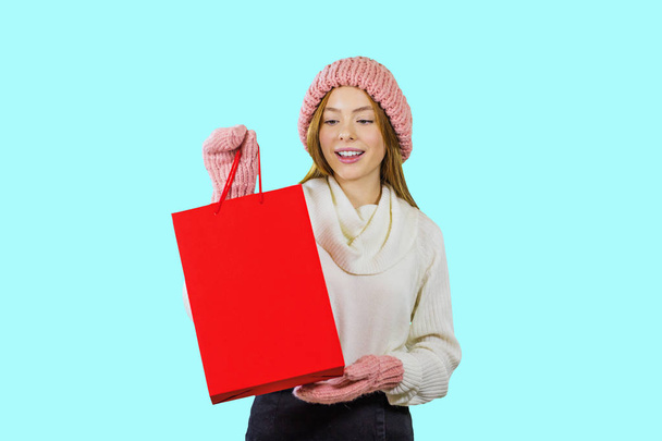 Portrait of a cute red-haired girl in a knitted hat and mittens holding a red bag and looking at it showing something on an isolated background, fashion shooting, New Years gifts - Zdjęcie, obraz