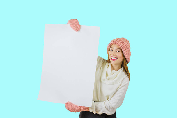 A young red-haired girl in a knitted pink hat and mittens is holding a large poster of white color with both hands and looking out at him smiling at the camera. - Photo, Image