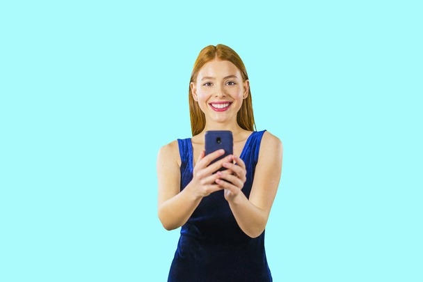 Portrait of a beautiful young red-haired girl in a blue velvet dress with a phone in her hands who is looking at the camera and smiling - Фото, зображення