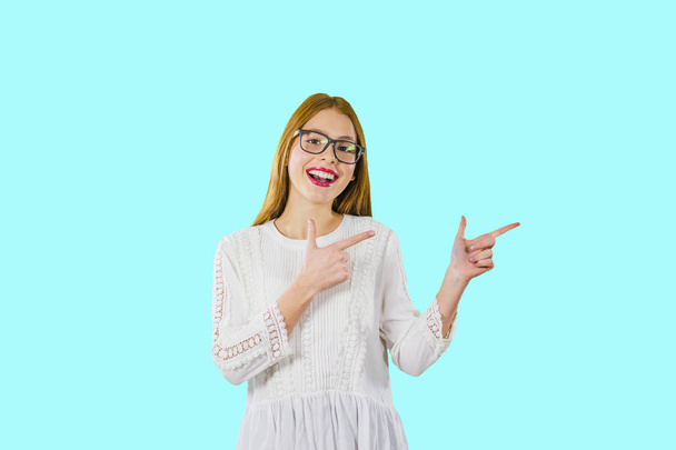 A portrait of a young beautiful girl with long red hair with glasses who shows with the index fingers of both hands to the side demonstrating something - Foto, imagen
