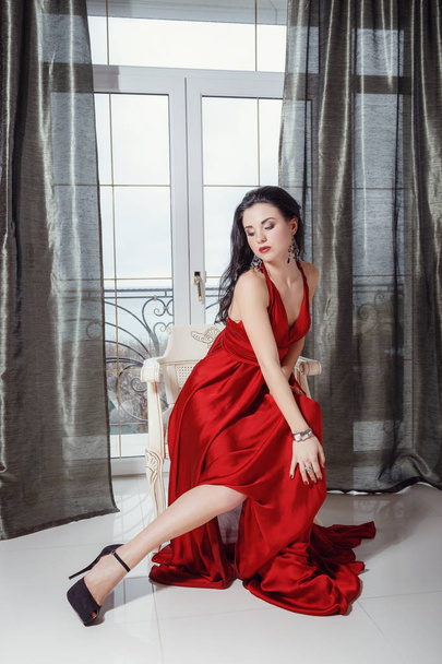seksowna kobieta Glamour z czarnymi włosami w eleganckiej czerwonej sukni siedzącej na fotelu - Zdjęcie, obraz