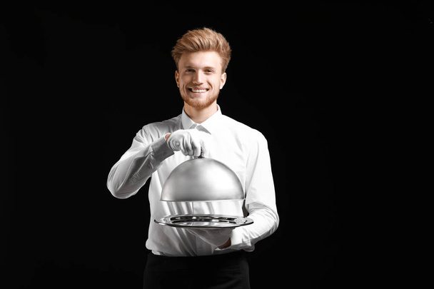 Handsome waiter with tray and cloche on dark background - Photo, Image