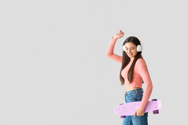 Dancing young woman with skateboard and headphones on light background - Photo, Image