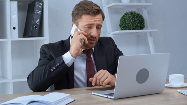 mature businessman using laptop and talking on smartphone in office - Materiał filmowy, wideo