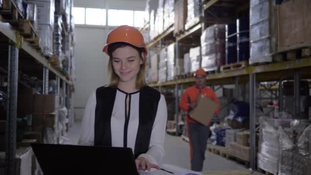 workwoman in helm praat met magazijnmedewerker tijdens het gebruik van laptop tussen rijen racks - Video