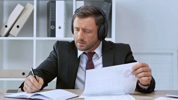 dospělý obchodník pracující s dokumenty a naslouchající hudbě na pracovišti - Záběry, video