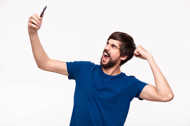 Knappe Brunet bebaarde man met snorren maken selfie met een smartphone tonen spier staande geïsoleerd over witte achtergrond. - Foto, afbeelding