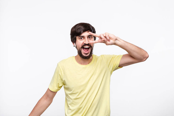 Excited brunet bearded man in a yellow t-shirt  with moustaches enjoying music listening songs with wireless headphones and dancing isolated over white background. - Foto, Bild