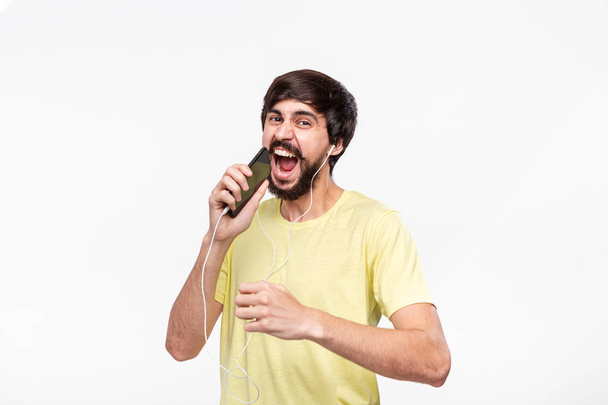 Handsome brunet bearded man with whiskers in a yellow t-shirt singing and dancing enjoying music with spartphone and wired headphones. - Foto, Imagem