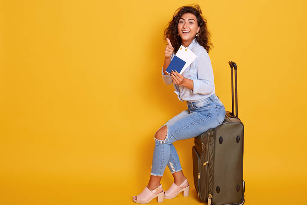Young brunette curly woman sits on grey luggage bag in front of yellow background pointing with her index finger at ticket in her hand. Female wearing stylish clothing, expresses happyness. Traveling - Photo, image
