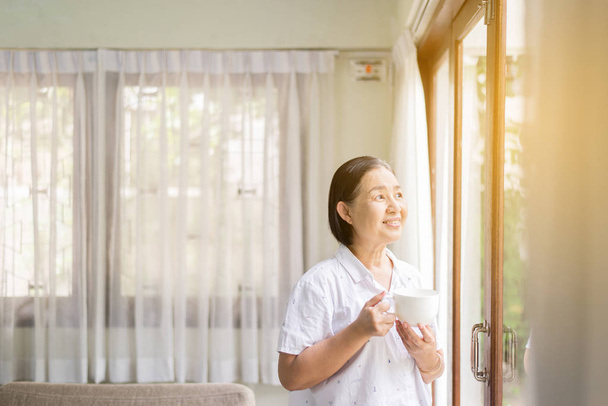 Senior asian woman holding a cup of coffee near the window in the morning,Happy and smiling,Positive thinking - Photo, Image