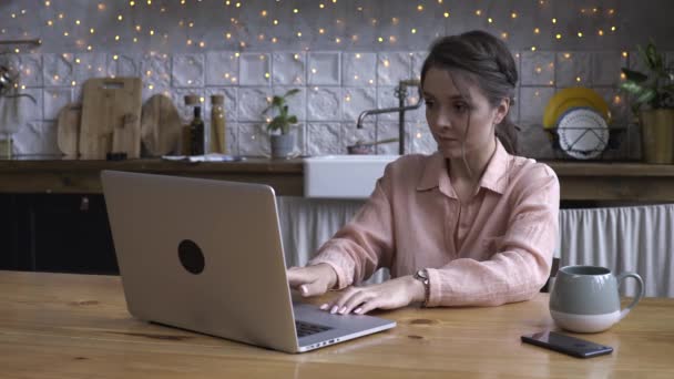 Close-up of attractive young young woman working on laptop computer while sitting on a kitchen. Stock footage. Modern decorated home interior on the background - Filmati, video
