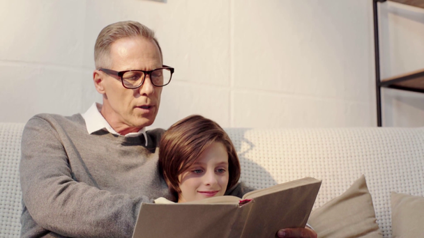 おじいちゃんと笑顔の孫がリビングルームで一緒に本を読む - 映像、動画