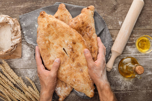 cropped view of man holding lavash bread on gray towel near rolling pin, flour package, wheat spikes and olive oil on wooden table - Photo, Image