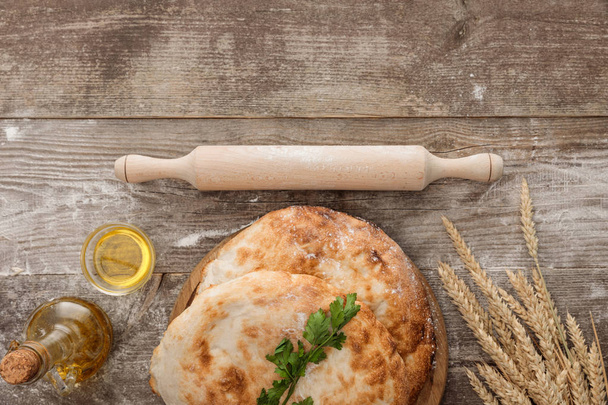 top view of lavash bread near wheat spikes, rolling pin, fresh parsley and bottle of olive oil on wooden table - Zdjęcie, obraz