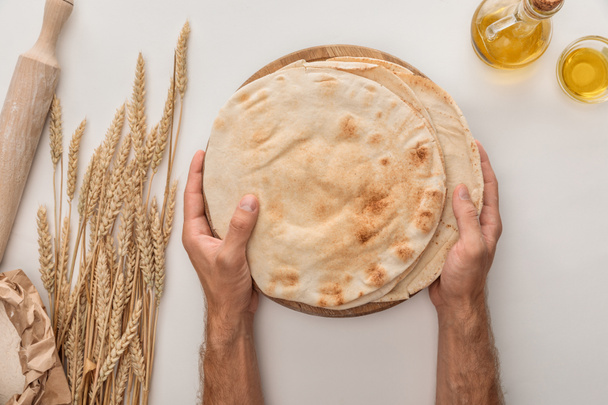 обрезанный вид человека, держащего плоский хлеб лаваш рядом с шипами пшеницы, скалка и оливковое масло на белой поверхности
 - Фото, изображение