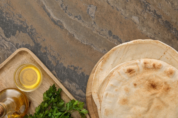 top view of flat lavash bread near wooden dish with parsley and olive oil on brown marble surface - Photo, Image