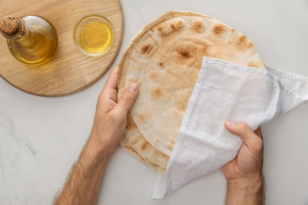 обрезанный вид человека, держащего плоский хлеб лаваш, покрытый белым полотенцем, рядом с растительным маслом на мраморной поверхности
 - Фото, изображение