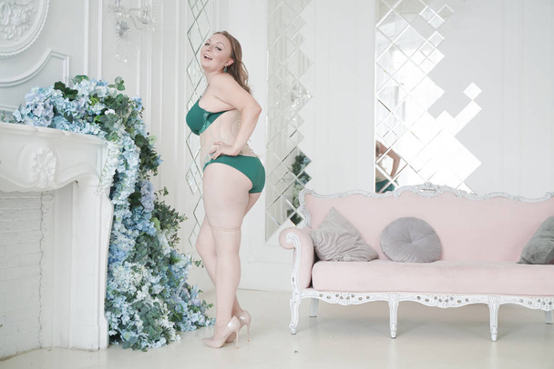 Портрет счастливой веселой толстой женщины в зеленом модном нижнем белье в белой комнате в помещении
 - Фото, изображение
