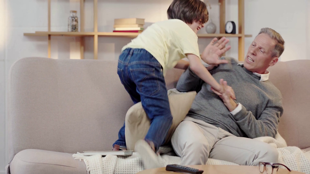 naughty grandson hitting granddad with pillow and jumping on sofa - Video, Çekim