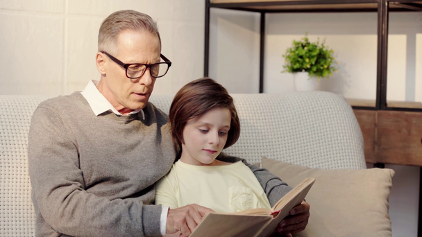 grandfather in glasses and grandson reading book together in living room - Filmati, video