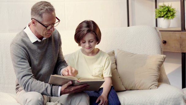 grandfather in glasses and grandson reading book together in living room - Filmati, video