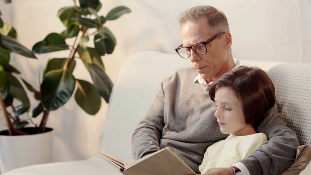 grandfather in glasses and grandson reading book together in living room - Metraje, vídeo