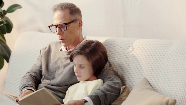 παππούς σε γυαλιά και ο εγγονός διαβάζοντας βιβλίο μαζί στο σαλόνι - Πλάνα, βίντεο