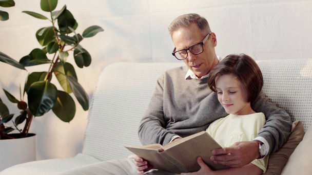 παππούς σε γυαλιά και ο εγγονός διαβάζοντας βιβλίο μαζί στον καναπέ - Πλάνα, βίντεο