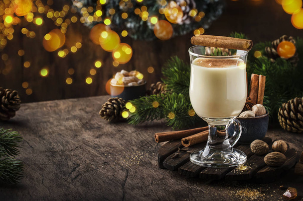 New Year or Christmas Eggnog cocktail - hot winter or autumn drink with milk, eggs and dark rum, sprinkled with cinnamon and nutmeg in a glass on wooden background, festive decoration - Photo, Image