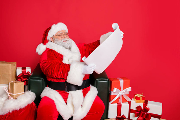 Portrait of his he nice cheerful kind generous fat bearded man St Nicholas reading address list clients wish want fairy miracle orders winter december isolated over bright vivid shine red background - Fotó, kép