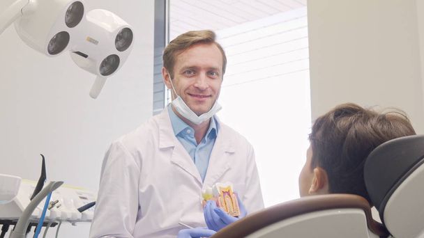 Alegre dentista masculino maduro sonriendo a la cámara mientras educa a un pequeño paciente
 - Foto, imagen