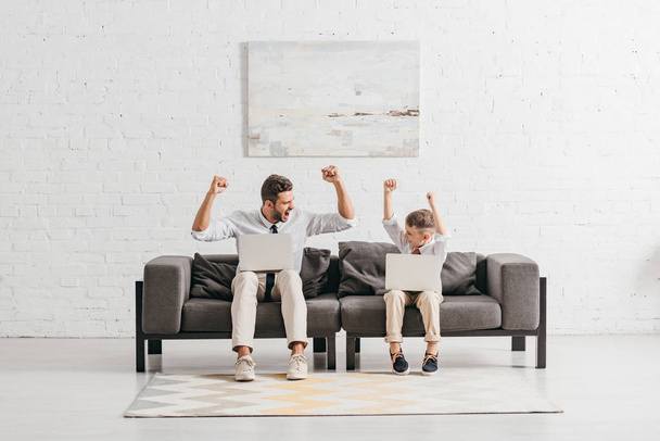vader en zoon in formele slijtage met behulp van laptops en tonen ja gebaren terwijl zittend op de Bank - Foto, afbeelding