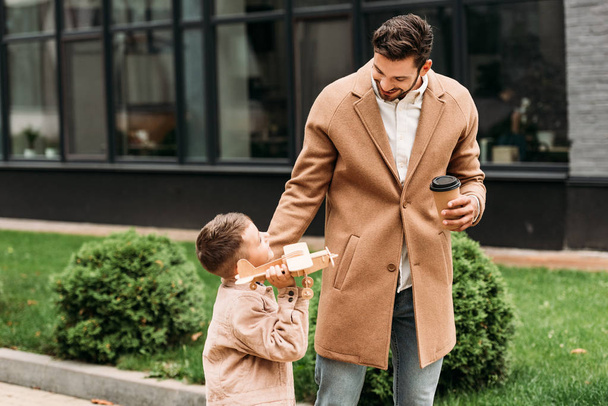 smiling father holding disposable cup of coffee and son with toy plane on street - Fotografie, Obrázek