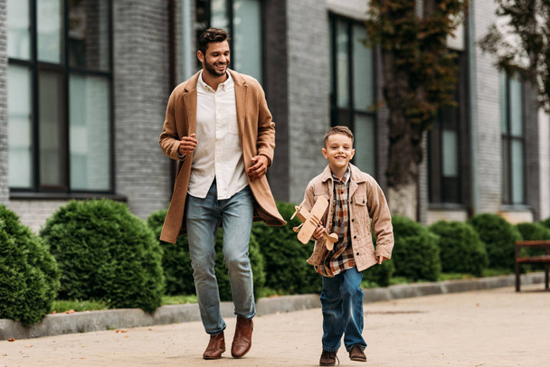 pohled na tátu v kabátě a synovi s hračkářovou rovinou běžící na ulici - Fotografie, Obrázek