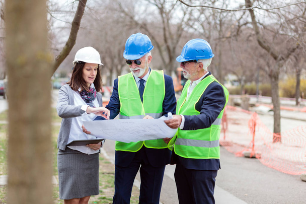 Smíšený tým starších mužů a mladých ženských architektů nebo obchodních partnerů, kteří se dívají na stavební plány na staveništi - Fotografie, Obrázek