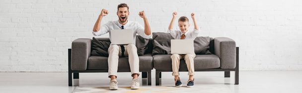 panoramic shot of smiling dad and son sitting on sofa with laptops and showing yes gestures - 写真・画像