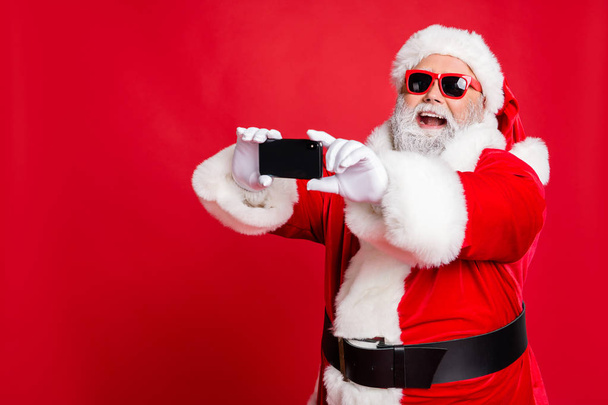Portrait of excited santa claus in eyewear making selfie with cell phone device on christmas vacation smiling wearing belt isolated over red background - Foto, Imagen