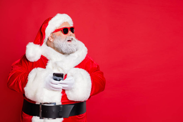 Portrait of cheerful santa claus in hat looking dreamy using cell phone plan newyear party wearing belt eyewear eyeglasses isolated over red background - 写真・画像
