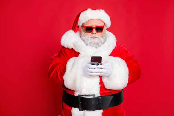 Portrait of minded santa claus with eyewear eyeglasses using cell phone device reading news wearing costume isolated over red background - 写真・画像