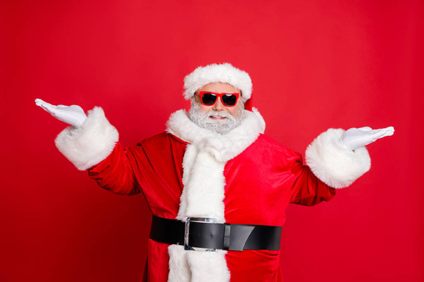Portrait of handsome cool funny santa claus with eyeglasses holding newyear ads promo wearing black belt cap isolated over red background - Fotó, kép