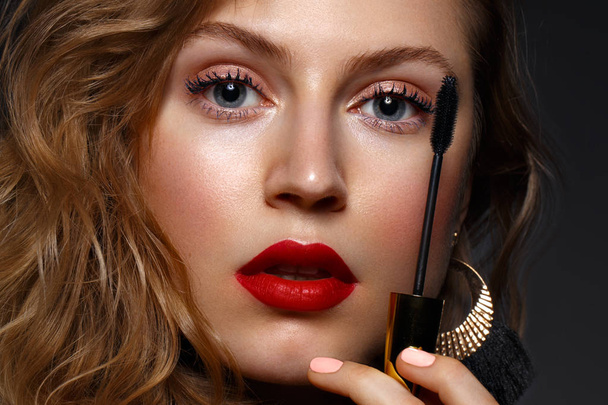 Beautiful girl with red lips and classic makeup and curls with mascara in hand. Beauty face - Φωτογραφία, εικόνα