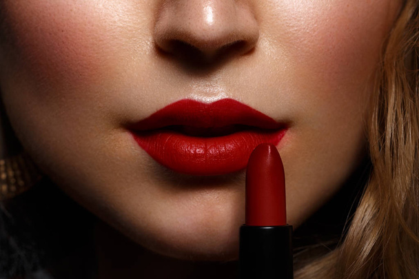 Mooi meisje met rode lippen en klassieke make-up en krullen met lippenstift in de hand. Schoonheid gezicht - Foto, afbeelding