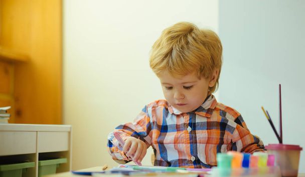 Kleiner Junge beim Zeichenunterricht. Kinder malen gerne mit Pinsel und Farben und tragen ein buntes Hemd. Kleine Kinder üben motorische Fähigkeiten, indem sie mit Feder zeichnen. Entwicklung von Aktivitäten für Kinder. - Foto, Bild