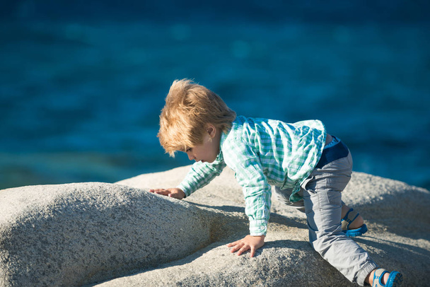 Kinderkletterstein. niedliche modische Baby Boy in blauem Hemd und grauer Hose klettert auf Stein an einem sonnigen Sommertag auf natürlichem Meeresgrund. Der kleine blonde Junge hat Spaß am Meer. Sport für Kinder. - Foto, Bild