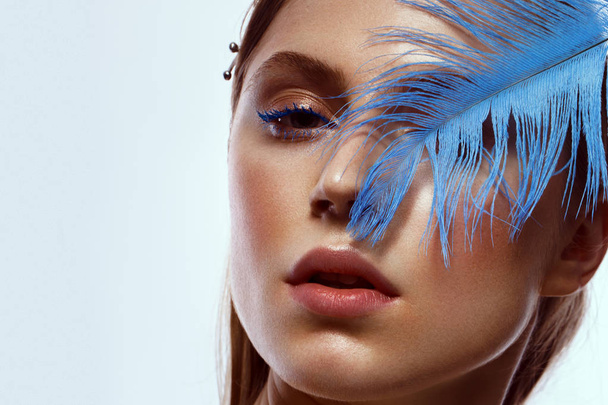Beautiful tanned girl with creative make-up and blue eyelashes, feather in hand. Beauty face. - Photo, Image