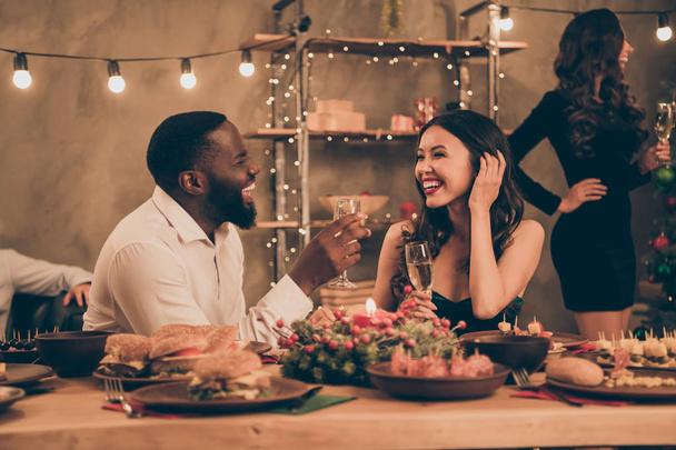 Profilseite Foto von fröhlichen Paar von gemischten Rassen Mann und chinesische Frau geben Toast Hold Champagner sprechen genießen Weihnachtsfeier x-mas night im Haus mit Tisch voller Neujahr Abendessen Kunststück drinnen - Foto, Bild