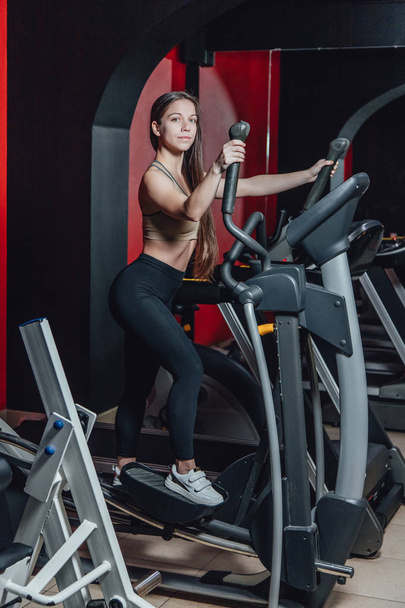 Portrait of a young and athletic woman running on a treadmill in a gym. Girl on the simulator, beautiful girl goes in for sports. - Valokuva, kuva